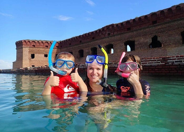 A mom holds both children, all wearing snorkel gear, with their heads out of the water.