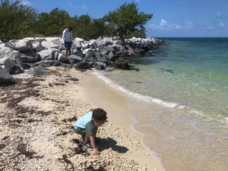 A young boy crouches to pick something up on the beach at Fort Zachary Taylor, while a his dad stand in the background admiring the ocean.