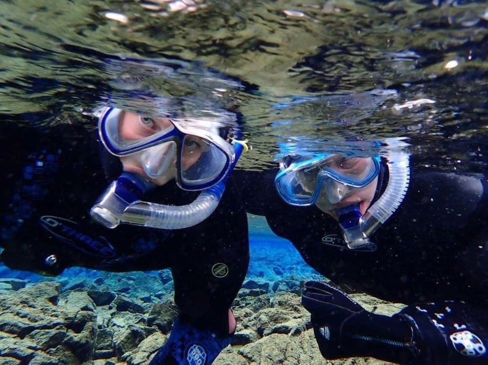 A close up of two snorkelers wearing black wet suits and blue goggles, snorkeling off the coast of Iceland.