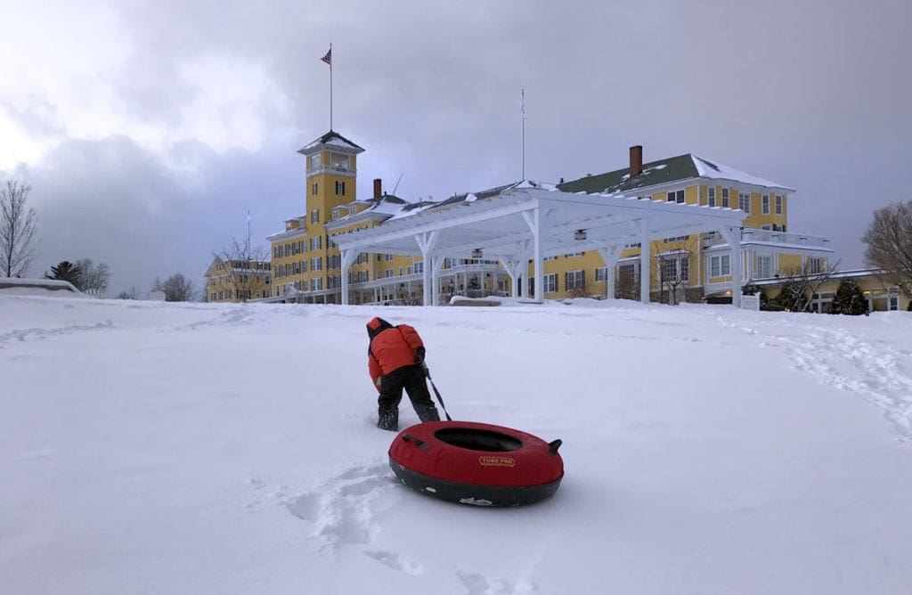 A young child wearing a red coat pulls a red tub sled up the hill toward the Mountain View Grand Resort and Spa, one of the best family resorts near NYC for a winter getaway.