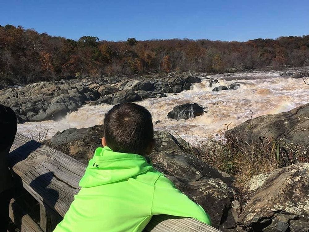 A young boy wearing a green hoodie leans over a rail looking out onto the river within Great Falls.