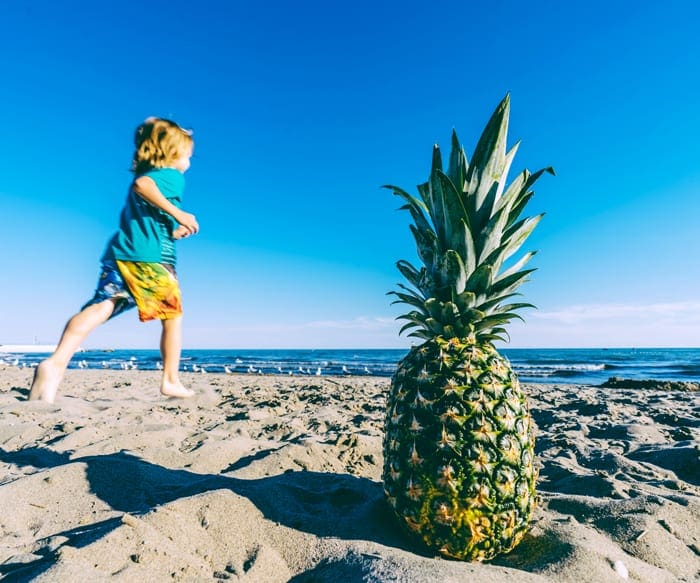 A young child runs along a beach, passed a pineapple resting in the sand.