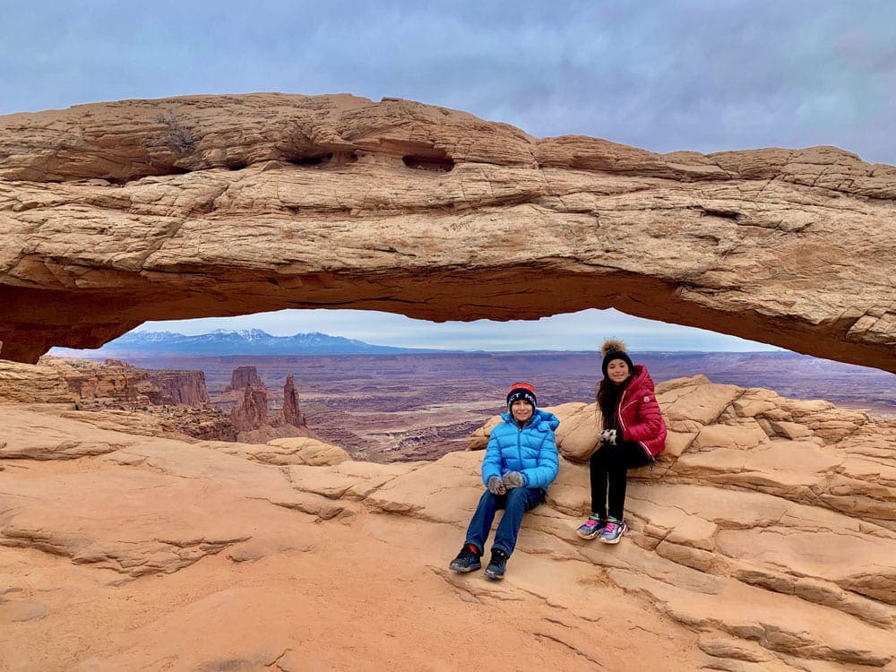 Two kids sit smiling near a low arch within Canyonland National Park.