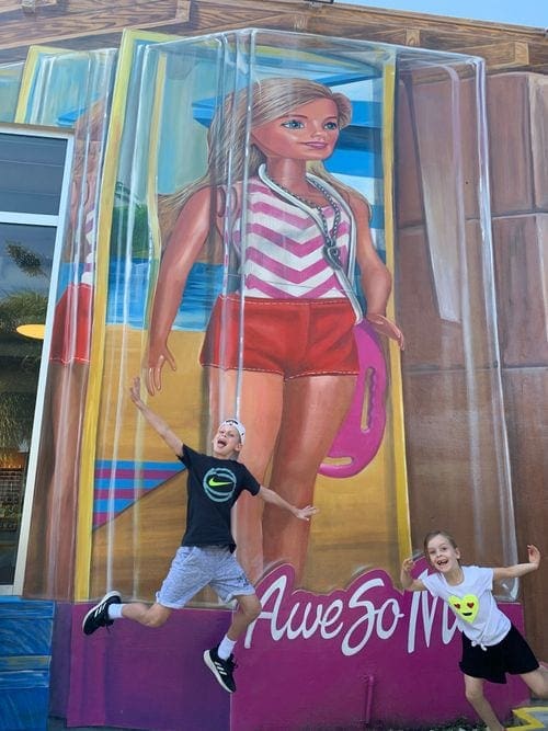 Two kids jump with silly faces in front of a colorful art installment featuring Barbie at Wynwood Walls in Miami.