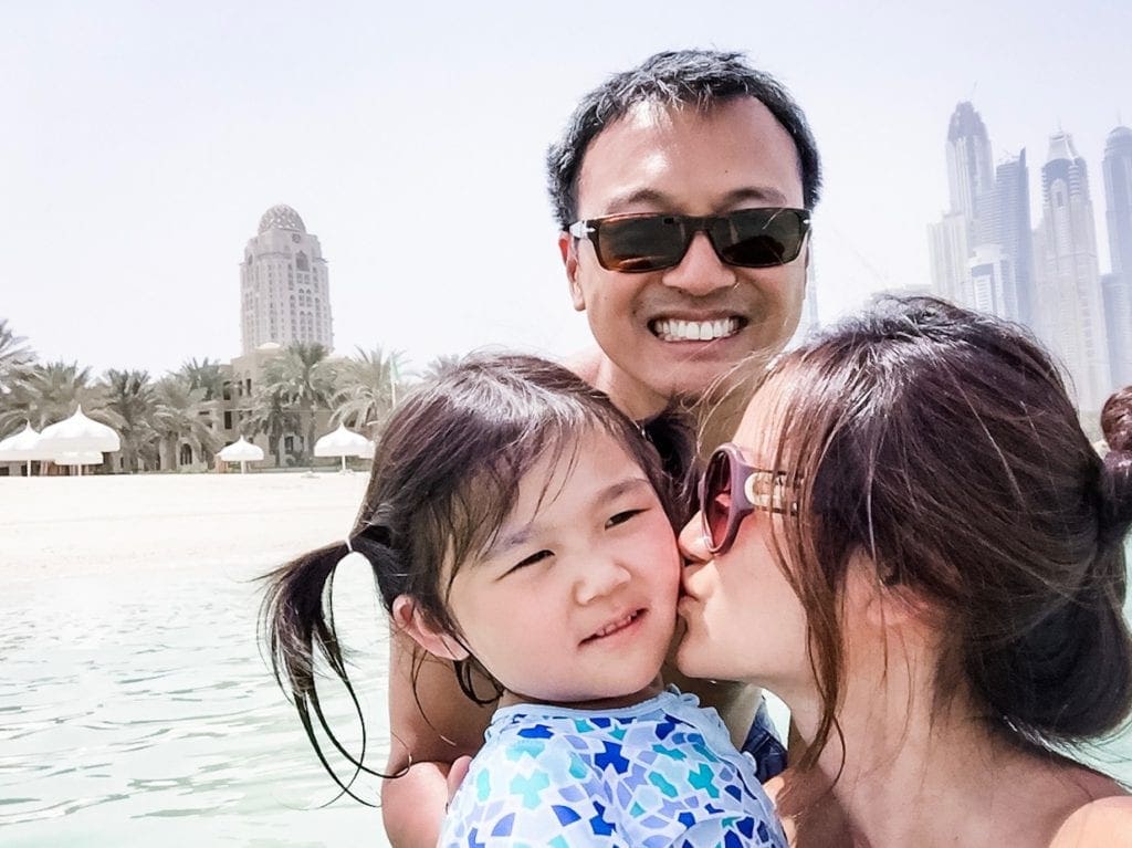 A mom kisses her young daughter while her husband smiles, the city skyline of Dubai is in the background.