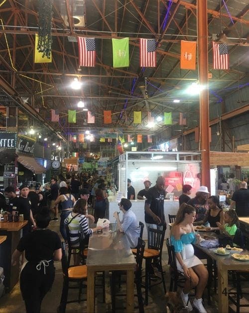 Inside a food court area within Little Havanna, featuring tables with people eating, flags, and food stalls.