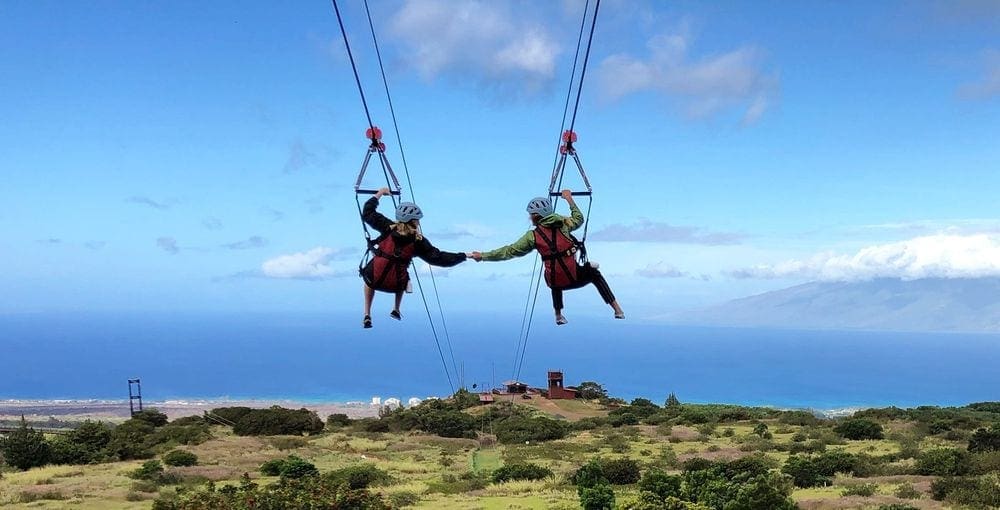 Two people zipline while holding hands with Kapalua Ziplines.