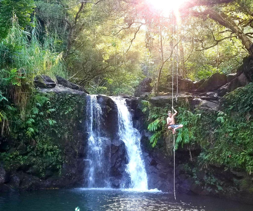 A young boy climbs a roap over a pool and waterfall in Maui, one of the best islands in Hawaii for kids.