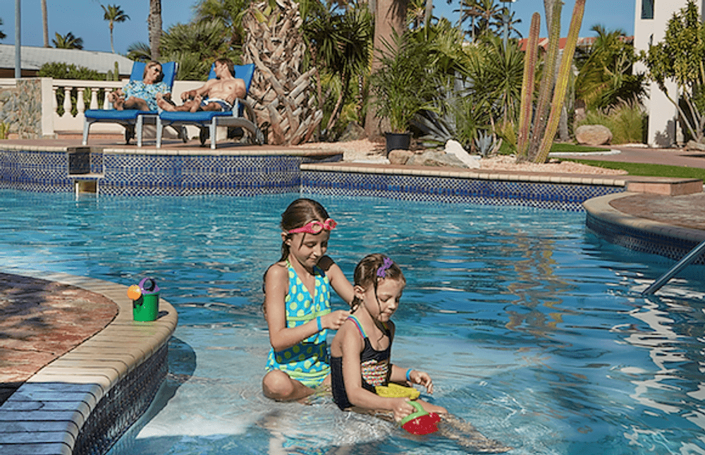 Two girls play in a pool while their parents watch from pool-side chairs at Tamarijn Aruba All Inclusives.