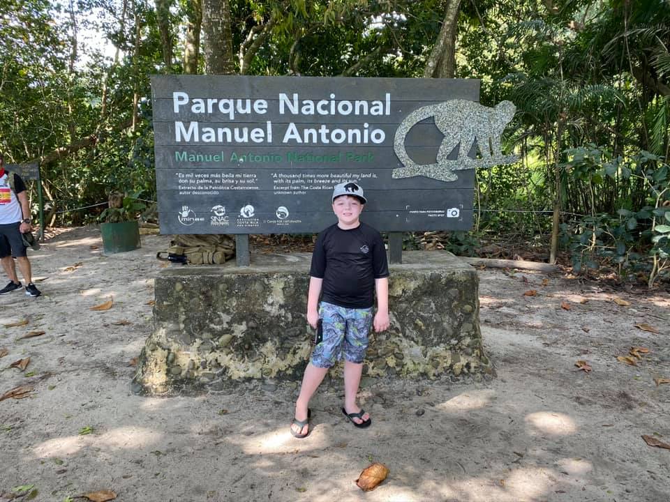 A young boy stands in front of the entrance sign for Manual Antonio National Park in Costa Rica.