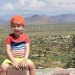 A young boy with a red cap on backwards sits on a stone fence with a desert view behind him while exploring Pinnacle Peak Park.