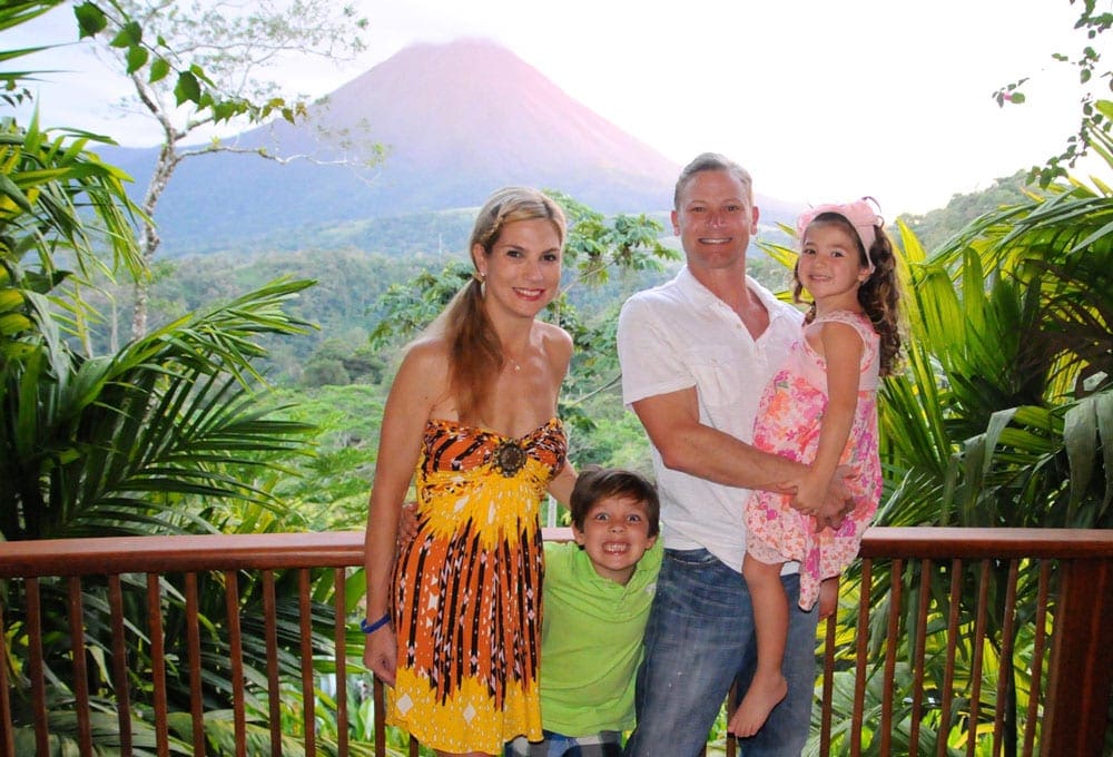 A beautiful family stands surrounded by lush foliage with Arenal Volcano in the background.