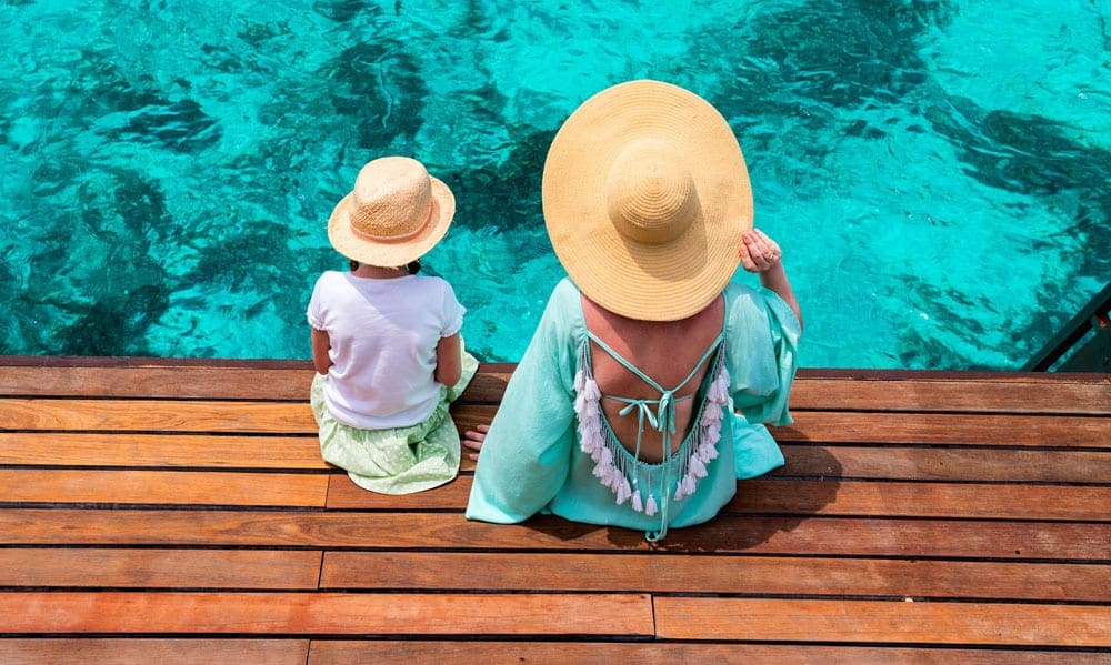 Mom and daughter sitting on a wooden platform of their over water bungalow watching the beautiful Maldives water, one of the best hot places to visit in December for families.
