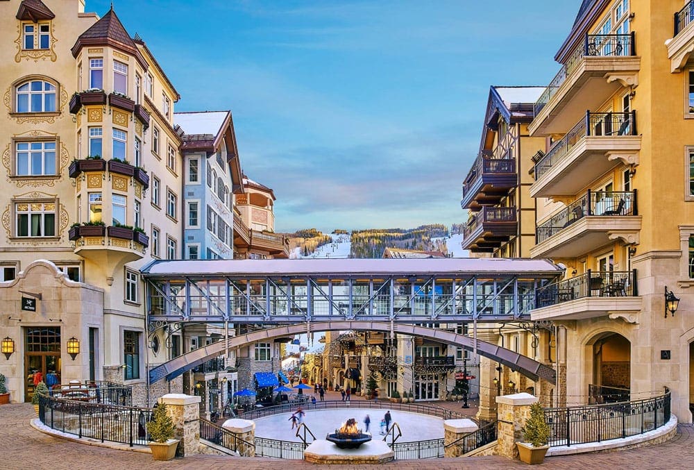 The skyway bridge resting between two resort buildings at the The Arrabelle at Vail Square, with a ice skating rink in the foreground.