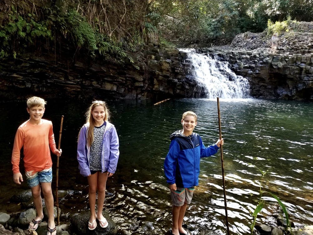 Three kids stand on rocks on the water while visiting Twin Falls in Hawaii, which is a great place to stop on the Road to Hana with kids.