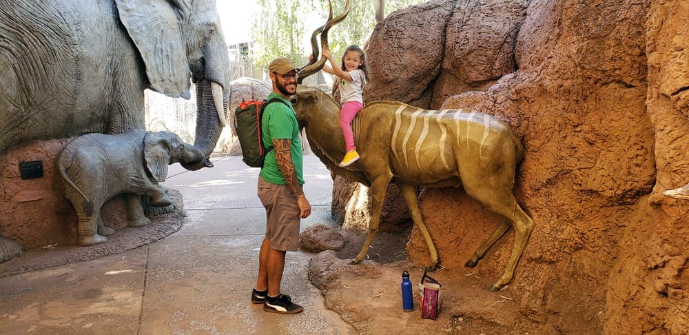 A dad stands near his young daughter as she rides a statue of an African animal at the Phoenix Zoo, one of the best things to do in Phoenix with kids!