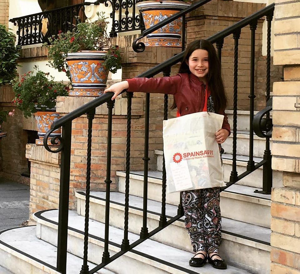 A young girl stands on a Seville sidewalk with a shopping bag across her shoulder.