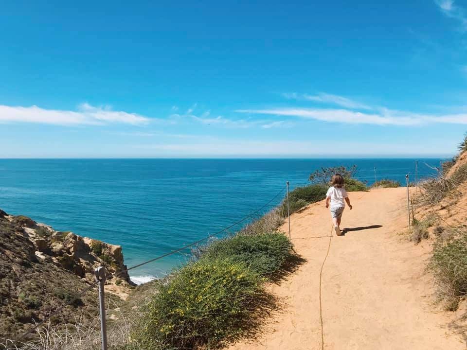 A young boy wanders a dirt path in San Diego, with the ocean beyond it, on a sunny day.