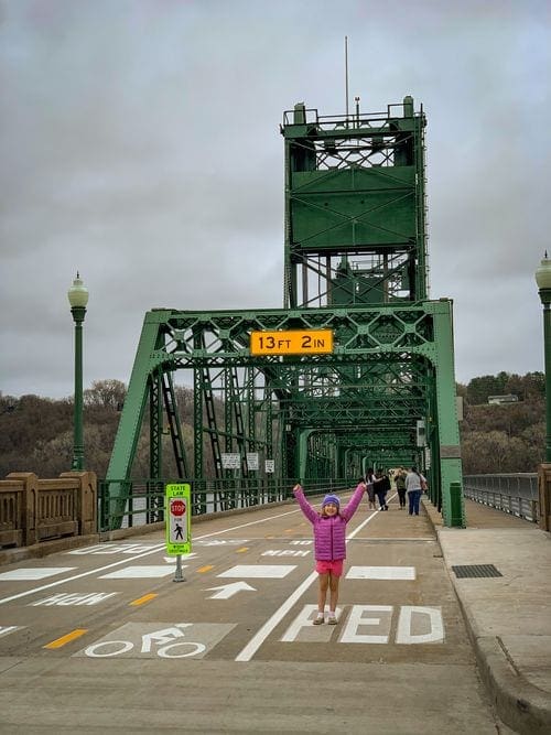 A young girl throws her hands in the air while exploring a pedestrian footbridge in Stillwater, Minnesota, a great stop for a Midwest road trip with kids.