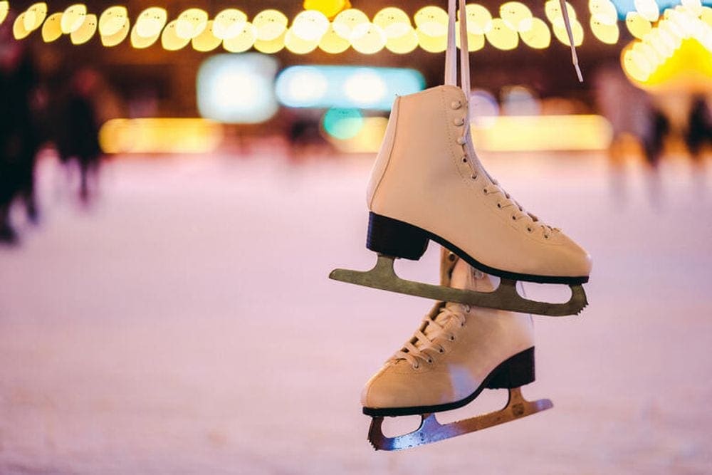 A pair of white ice skates dangles with a rink and lights behind them.