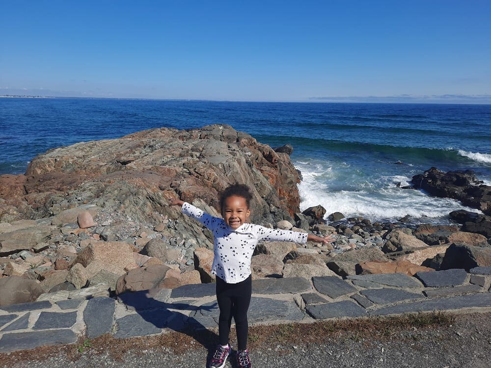 An African American toddler girls holds her arms wide open with a huge smile on her face, with Ogunquit Beach in Maine behind her, one of the best weekend getaways near Boston for families.