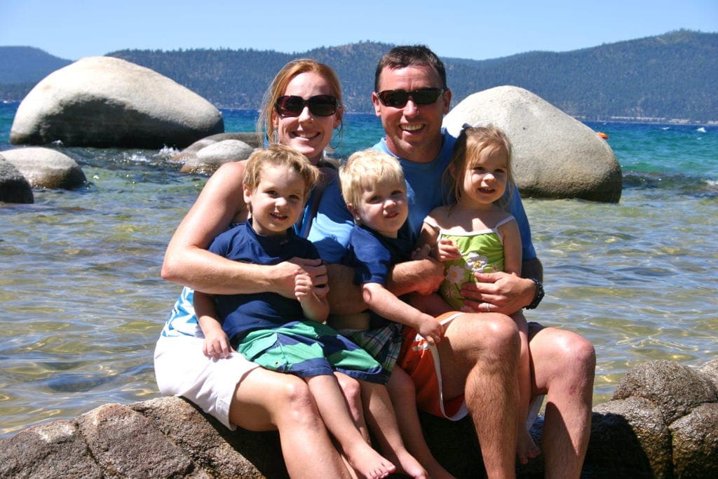 A mom and dad sit smiling with their three kids at Sand Harbor beach in Lake Tahoe, , one of the best Summer Vacation Ideas in the U.S. for Families. Large rocks nestle in the water behind them.