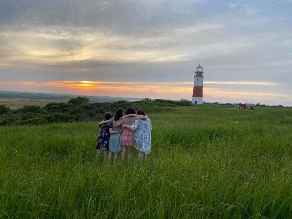 Four girls stand arm and arm admiring a sun set over a distant lighthouse in Nantucket.