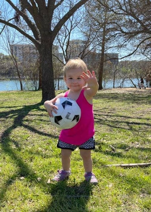 A young toddler holds a ball and waves on a sunny day at Lade Bird Lake in Austin.