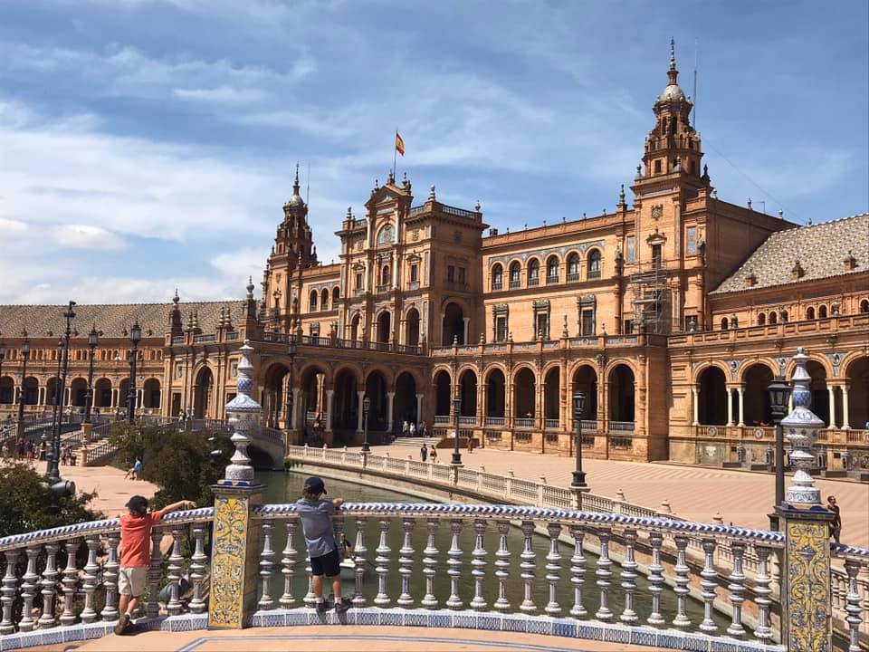 Two kids lean on a barrier facing a large Spanish building.