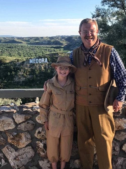 A costumed man and child stand with a view of Medora and its iconic Hollywood-esque sigh in the background.