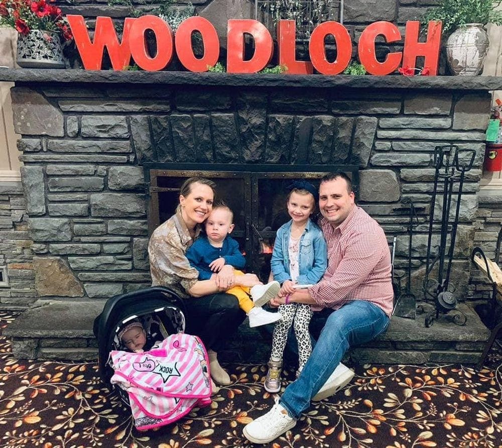 A family of five sits on the side of a fire place at Woodloch, with a large red sign overhead reading "Woodloch", one of the best resorts in Pennsylvania for families.