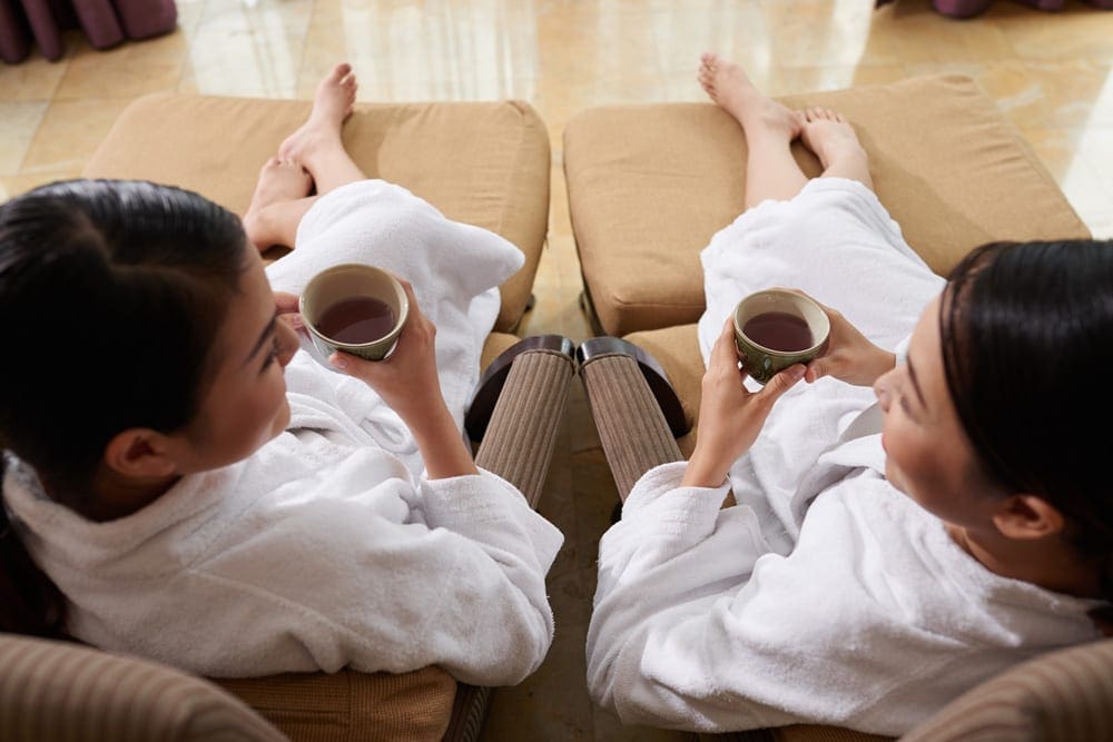 Two moms enjoy a cup of coffee while sitting in robes on spa loungers, a spa experience is one of the best family travel gifts of the year.