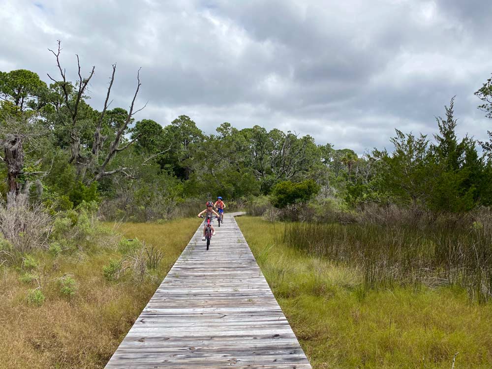 Two kids on bikes race down a boardwalk while exploring Kiawah Island, one of the best beaches in South Carolina for families. 