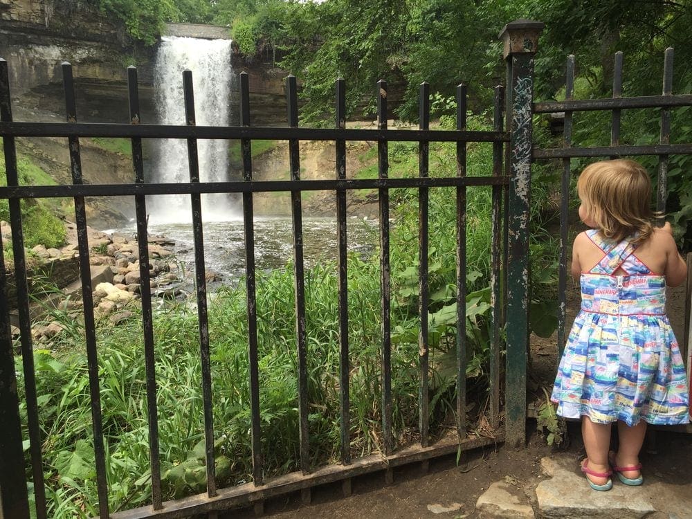 A young girl holds onto the railing while she peer at Minnehaha Falls in Minneapolis, one of the best stops in the Twin Cities with kids.