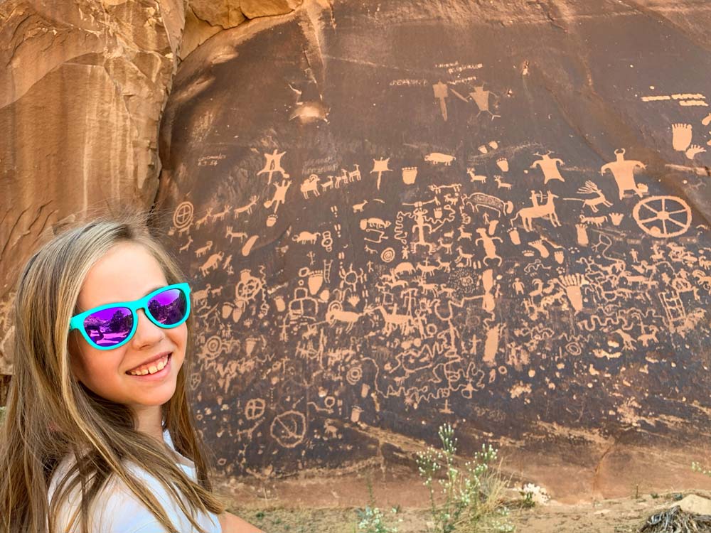 A young girl wearing sunglasses smiles at the camera while standing near the cave drawings inside Newspaper Rock, one of the best things to do in Moab with kids.
