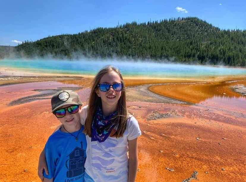 Two kids stand smiling with the Grand Prismatic behind them at Yellowstone National Park.