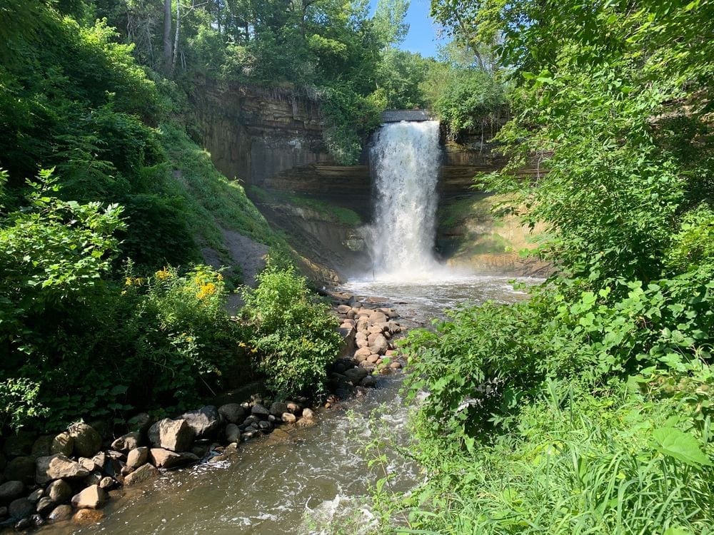 A large urban waterfall spills over the edge, surrounded by green foliage in Minneapolis.