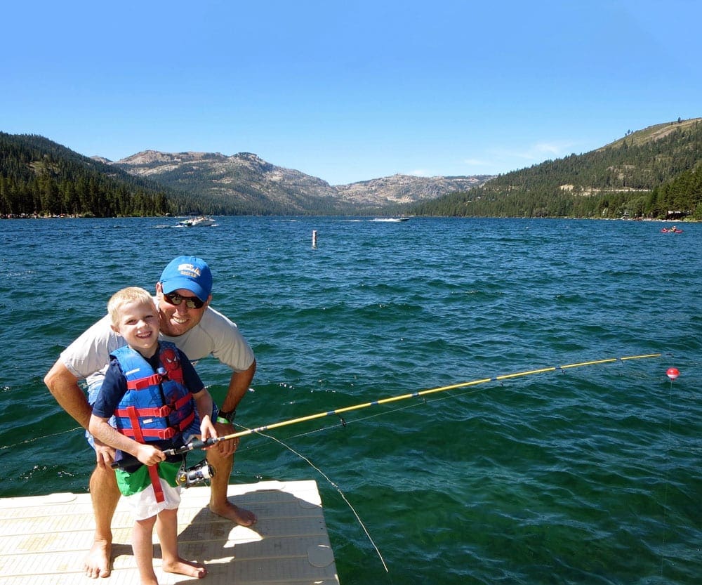 A boy holding a fishing pole smiles with his dad as they enjoy a beautiful day on Lake Tahoe, one of the top father-son weekend ideas.