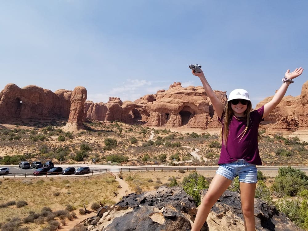 A young girls excitedly throws her hands in the air with the iconic Utah red rock formations in the distance.