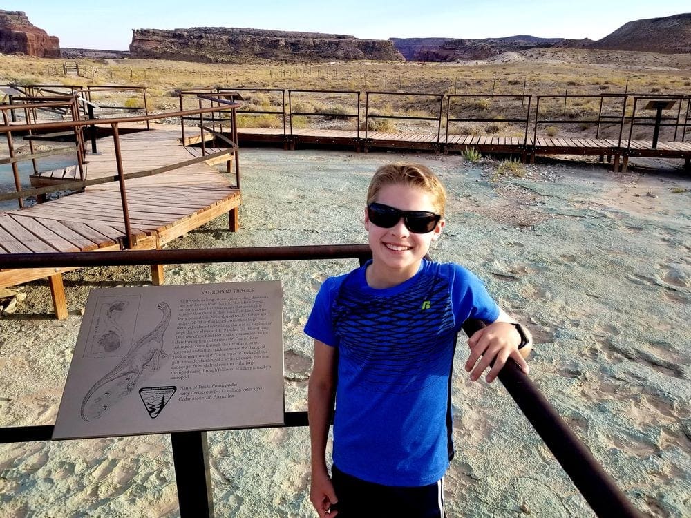 A young boy wearing a blue shirt and sunglasses stands near a sign featuring a dinosaur, while wandering the Dinosaur Trail in Utah.
