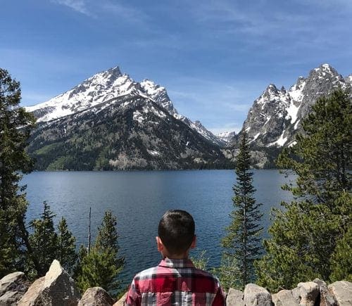 A young boy looks out over a lake with the Grand Tetons in the distance across a lake.