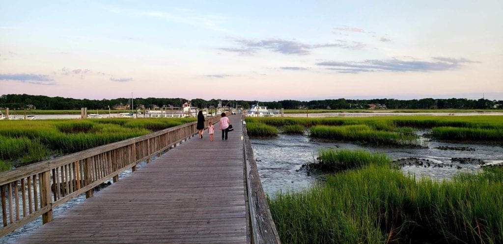 Three generations of women walk down a boardwalk in Hilton Head, with grassy wetlands on both sides.