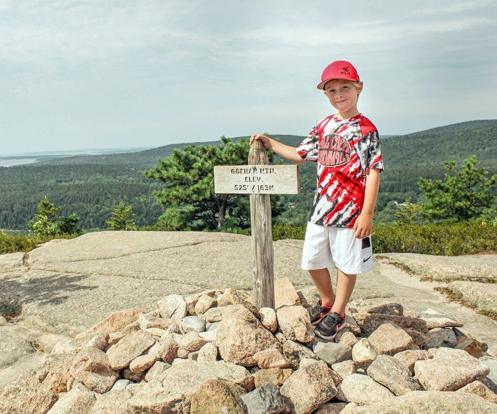 A young boy stands next to a summit sign on Bass Head Lighthouse Trail peek near Bar Harbor, one of the best East Coast summer destinations for families.