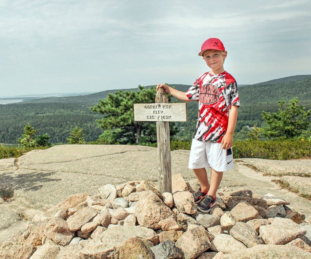 A young boy stands next to a summit sign on Bass Head Lighthouse Trail peek near Bar Harbor, one of the best places to visit on a Northeast road trip with kids. 