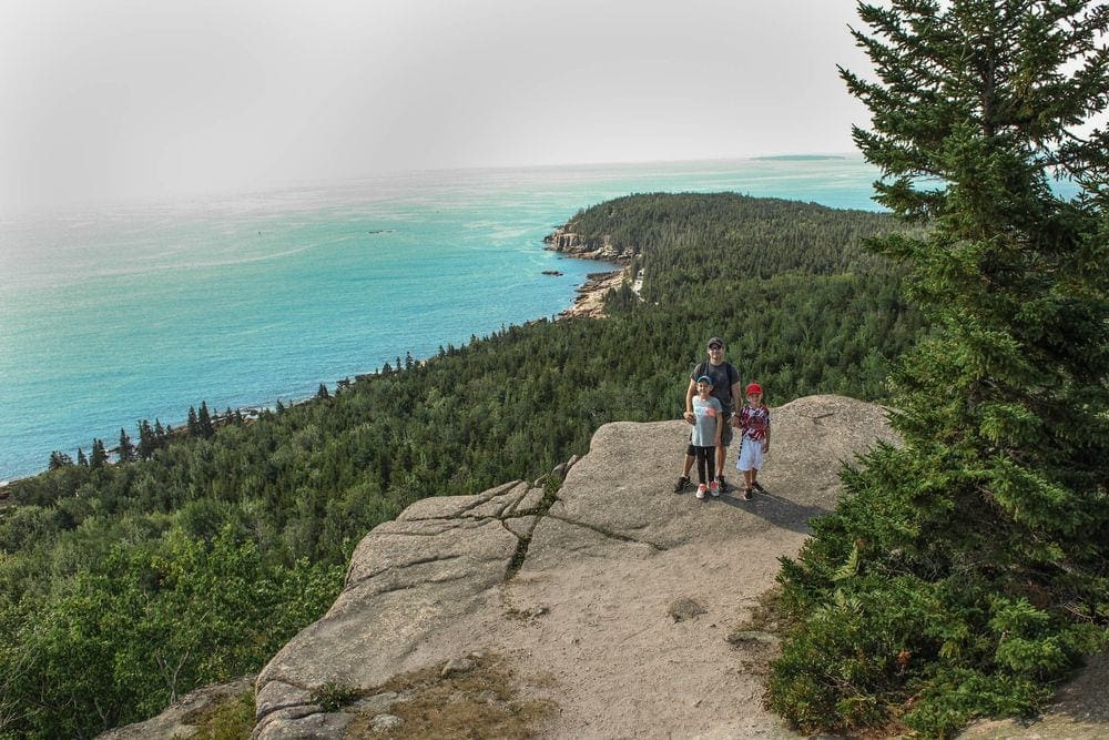A dad stands with his two kids atop a mountain cliff overlookng Acadia National Park in Bar Harbor, one of the best East Coast summer destinations for families.