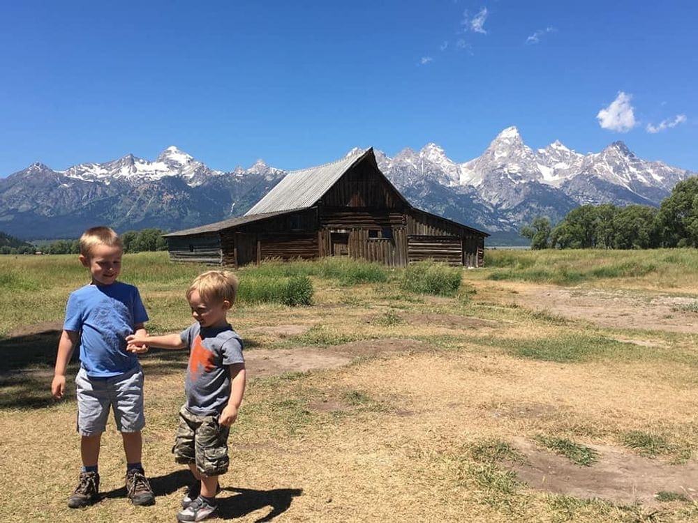 Two kids smile while walking together, holding hands, with a log building in the background and the Grand Tetons in the distance.