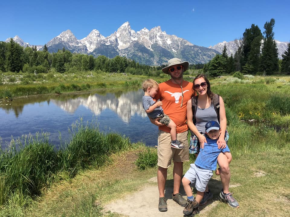 A family of four stands together smiling beside a glassy lake and the Grand Tetons in the distance.