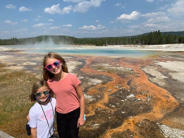 Two kids stand smiling with the Grand Pristmatic behind them at Yellowstone National Park.