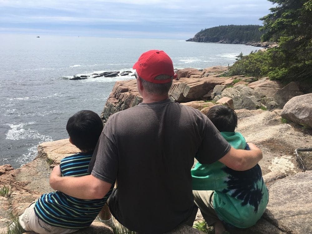 A dad holds his two young kids close while sitting atop a rock ledge in Acadia National Park, one of the top father-son weekend ideas.