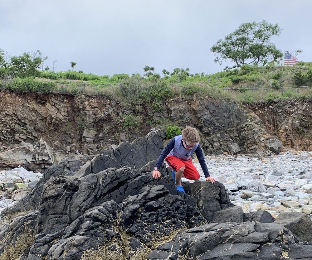 A young boy climbs around large rocks on the beach near Portland, Maine, one of the best weekend getaways near Boston for families.